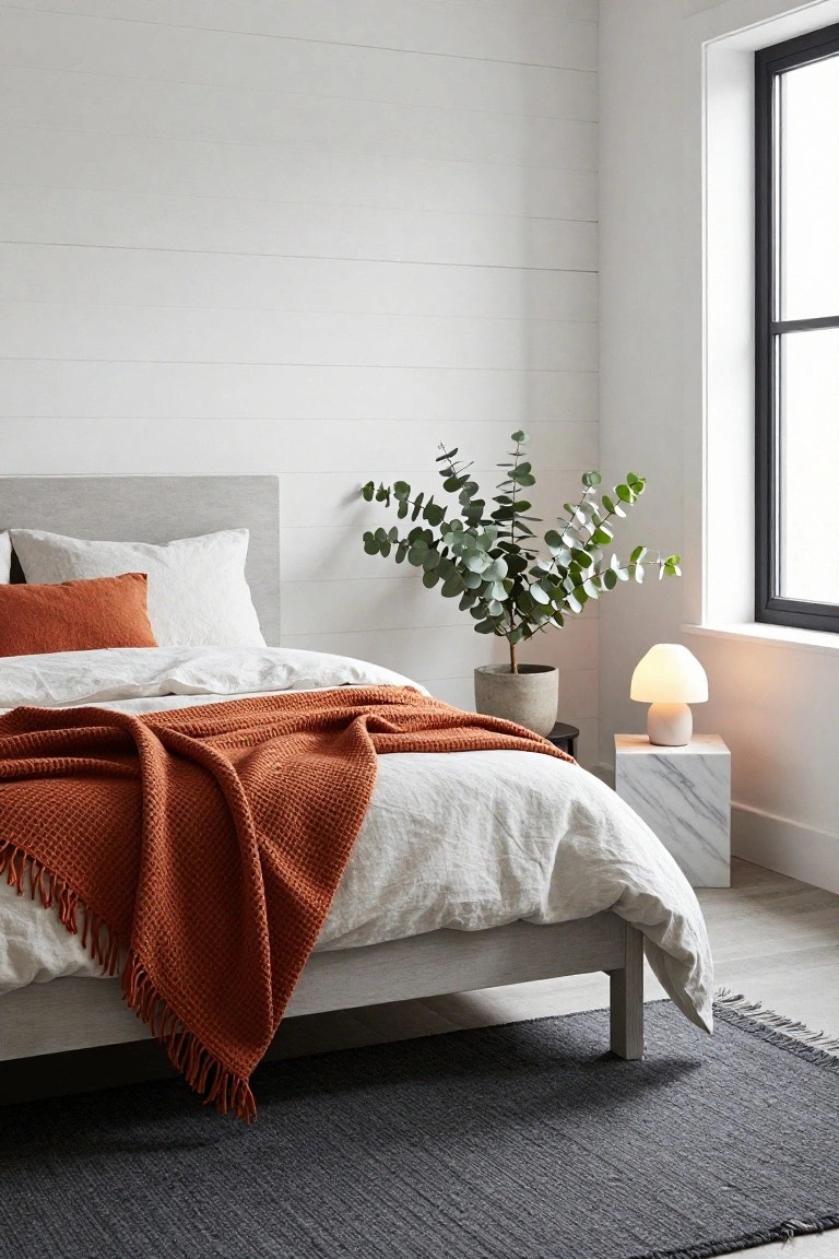 Bedroom corner showing a low-profile bed with gray headboard, white linen bedding, and draped orange chunky knit throw, next to a potted eucalyptus plant, white mushroom-shaped lamp on marble pedestal table, large black-framed window, white shiplap walls, and dark seagrass rug.