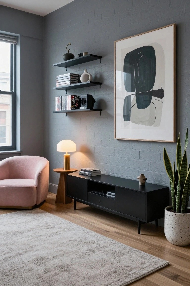 Interior corner of a room with gray brick accent wall, black floating shelves holding books and decor, large framed abstract black and white artwork, pink upholstered swivel armchair beside a round wooden side table with brass lamp, black media console with books and potted snake plant on light wood floors with beige rug.