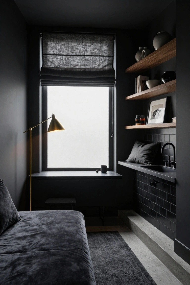 Small dark interior room with charcoal walls, black subway tile sink nook with faucet, floating wood shelves holding ceramics and books, gold arc floor lamp, low gray velvet bed, and window with shades.