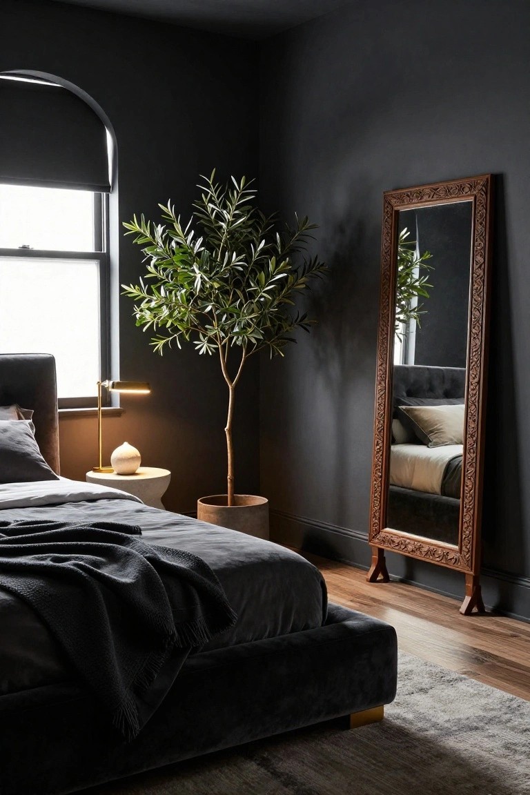 Dark charcoal painted bedroom with arched window covered by roller shade, velvet upholstered bed with gray sheets and throw, brass task lamp on side table with white vase, large potted olive tree next to tall carved wood full-length mirror, wood floor with gray rug.