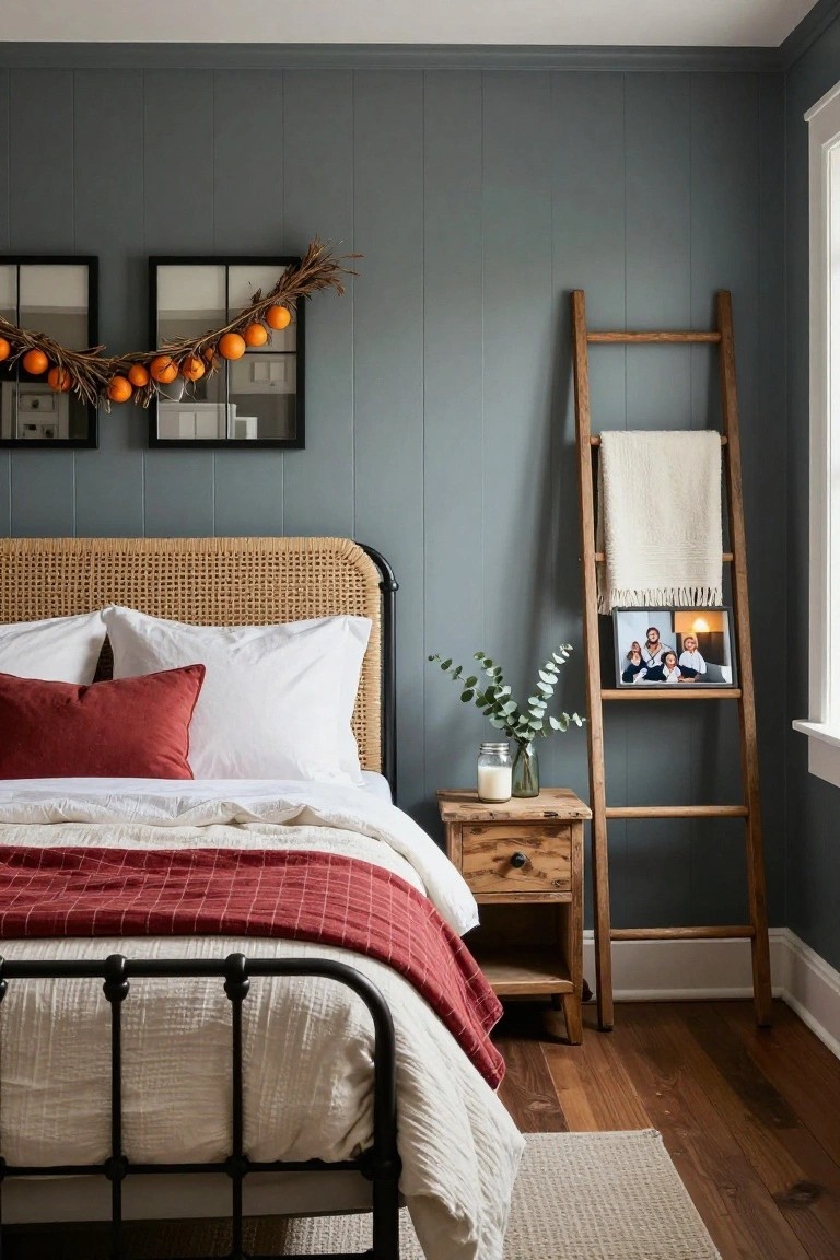 Cozy bedroom with gray shiplap walls, black metal bed frame with rattan headboard, white linens and red plaid throw, wooden ladder shelf draped with white blanket and holding family photo and eucalyptus jar, and dried branch garland with oranges over two black-framed mirrors.