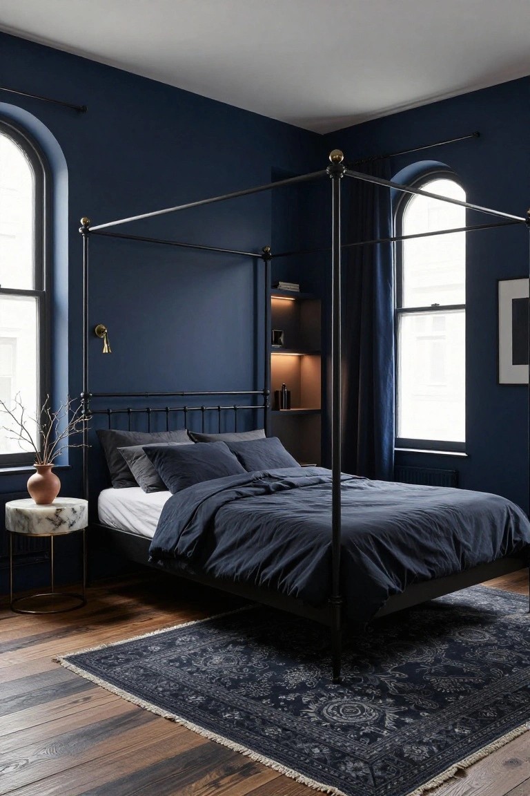 Corner of a bedroom with navy blue walls, black metal four-poster bed draped in navy bedding, marble side table with terracotta vase and branches, built-in shelves, arched windows with blue curtains, and navy Persian rug on hardwood floors.