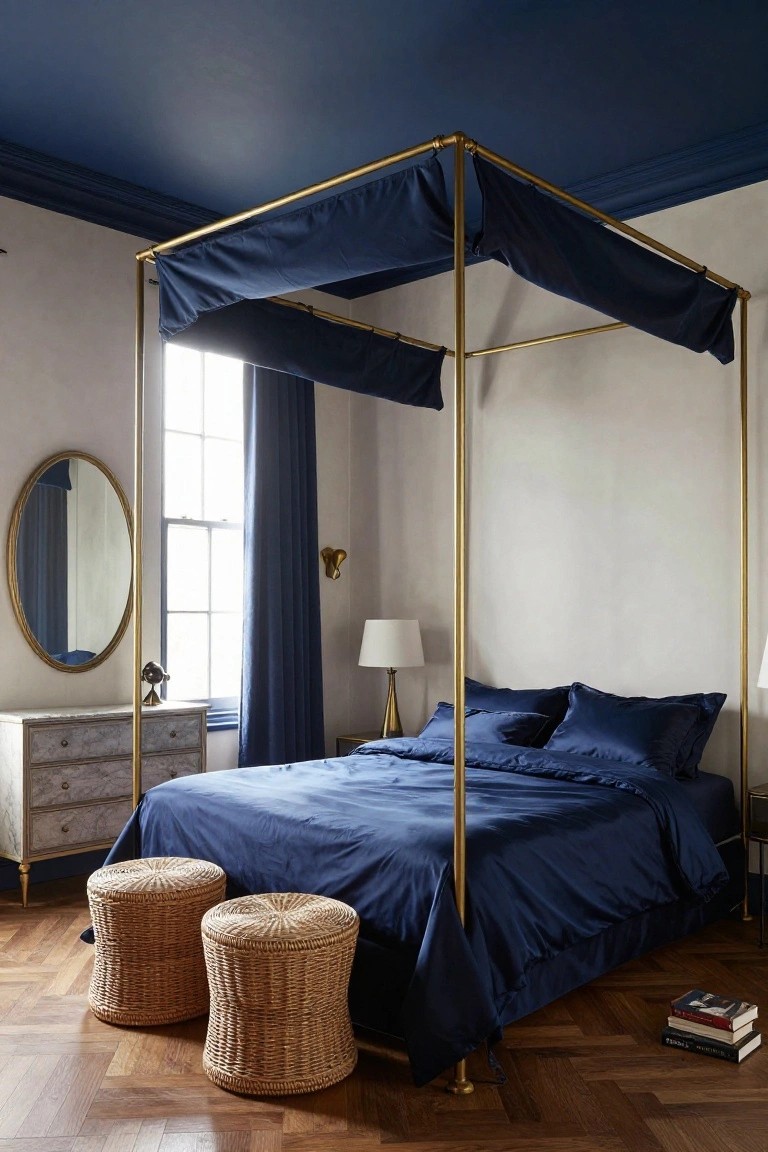 Bedroom with navy ceiling, brass four-poster bed draped in navy fabric, navy bedding and curtains, oval mirror, lamps, dresser, two wicker stools on parquet floor.