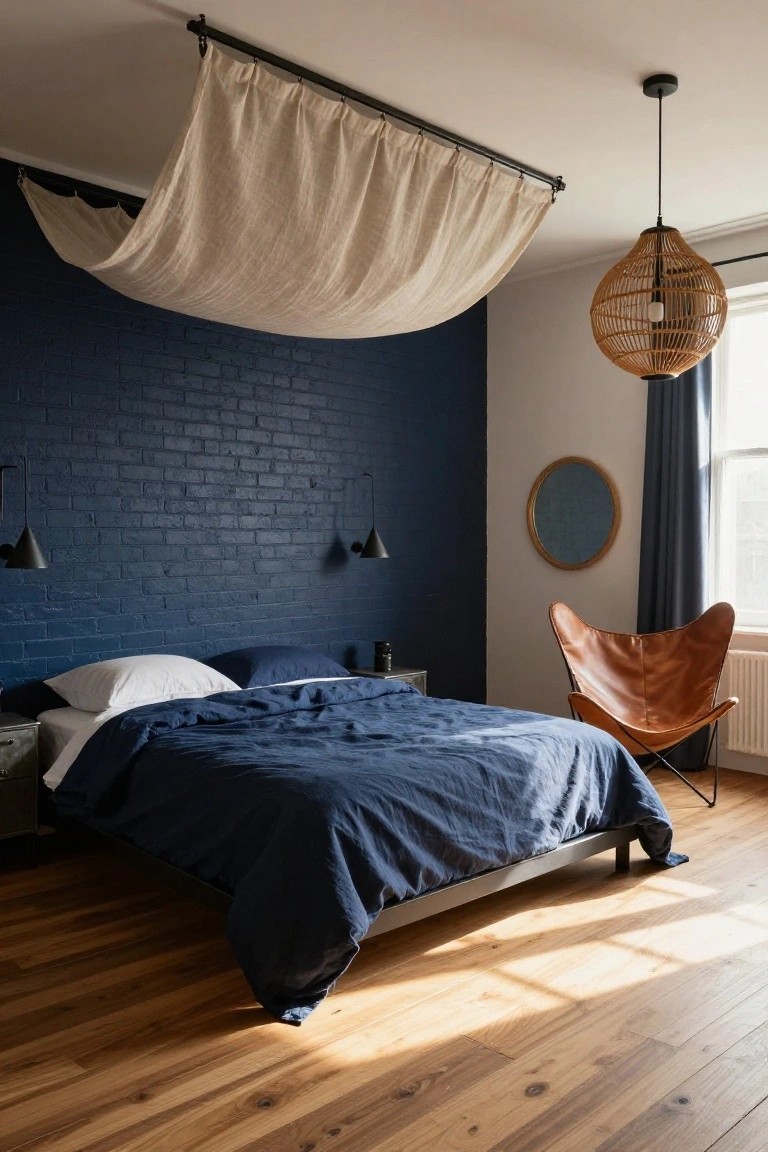 Bedroom featuring a navy painted brick accent wall behind a bed with navy linens and white pillows, a suspended white linen canopy overhead, black wall sconces, an orange leather Eames-style chair, rattan pendant light, and light wood floors.