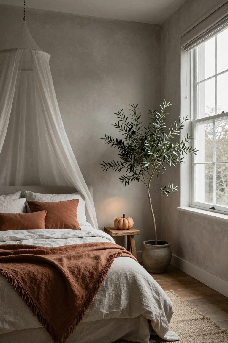 Bedroom corner with a low bed under a tall white sheer canopy, rust-colored pillows and fringed blanket, potted olive tree, small pumpkin on wooden stool next to large window, textured beige walls and wood floor.