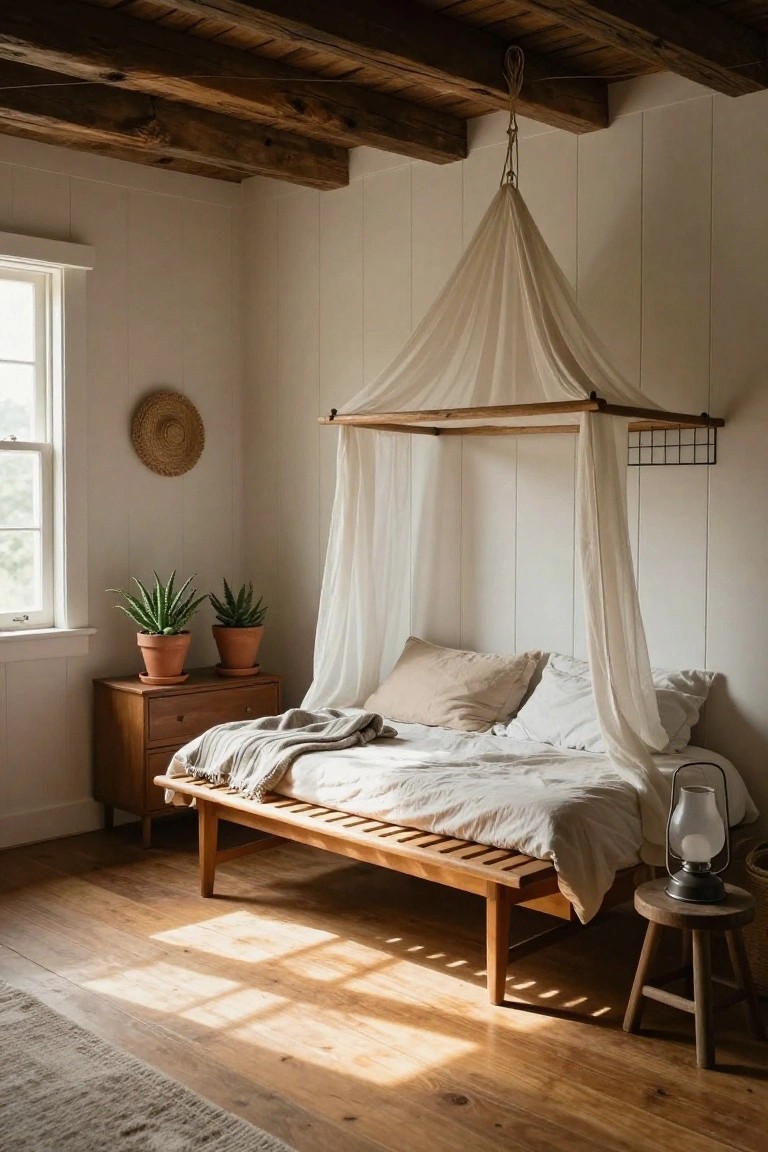 Sunlit bedroom with low wooden daybed under wooden canopy frame draped in sheer white fabric, two potted aloe plants on wooden dresser, small wooden stool holding metal lantern, white shiplap walls, wooden beam ceiling, hardwood floor, and woven rug.