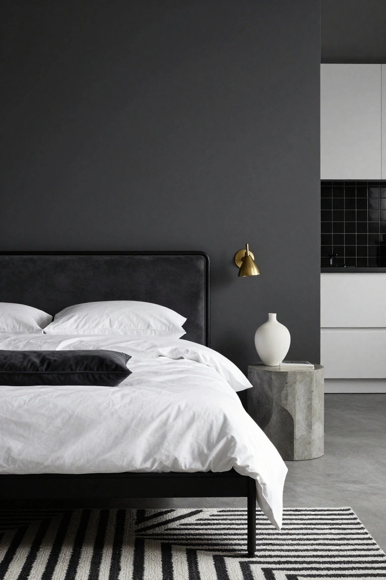 Modern studio bedroom featuring a black metal bed with dark gray velvet headboard and white bedding, gold wall lamp, concrete side table with white vase and book, dark charcoal wall, open white kitchen cabinets with black tiles, concrete floor, and black-and-white striped rug.