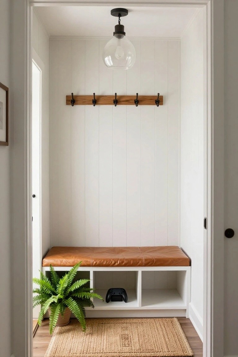 White shiplap-walled mudroom featuring wooden coat hooks mounted on the wall, a built-in white bench with tan leather cushion and open cubbies below holding shoes and a black gaming controller, a potted fern plant beside it, seagrass rug on wood floor, and glass pendant light overhead.
