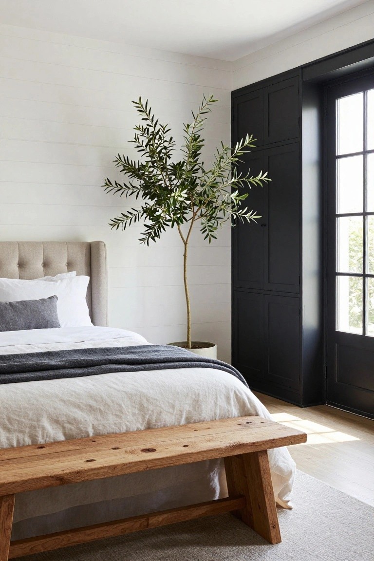 Bedroom interior featuring a beige tufted headboard bed with white sheets, gray throw, and pillows, a tall potted olive tree, black paneled wardrobe doors beside a multi-pane window, and a rustic wooden bench at the foot of the bed on light wood floors with a gray rug.