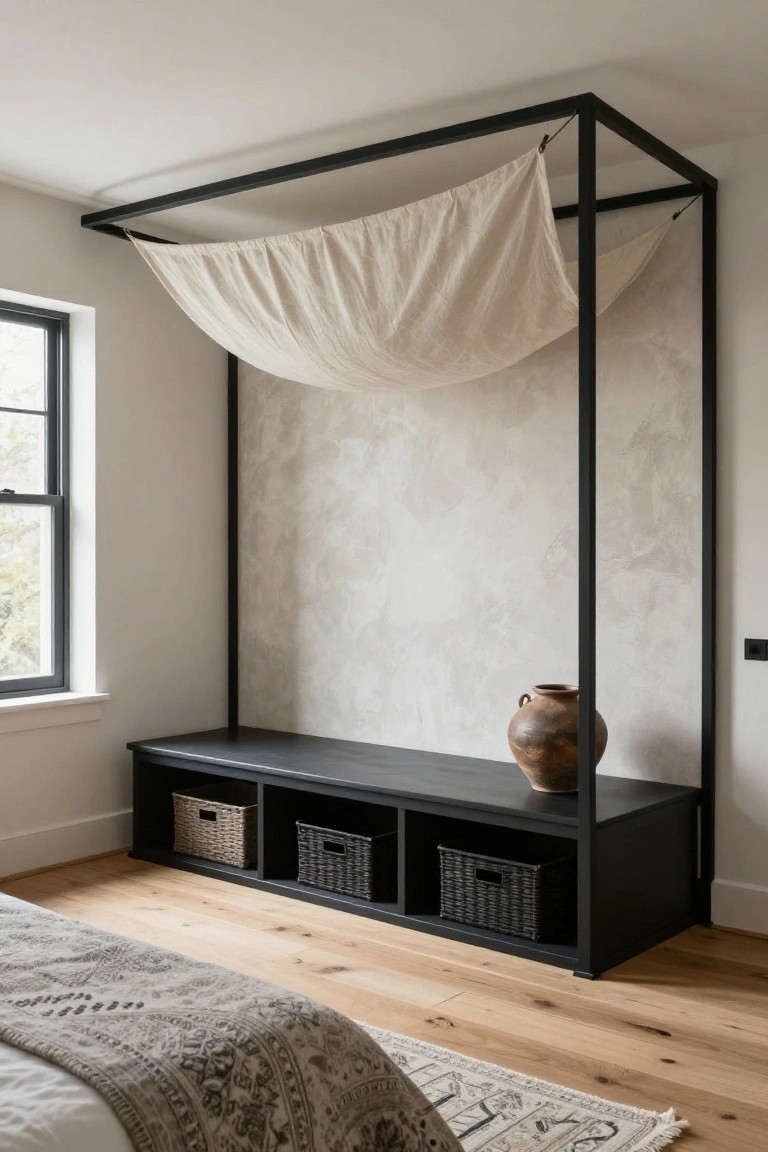 Black metal canopy frame with draped beige linen fabric over a black wooden bench with wicker baskets in a bedroom corner next to a bed with gray patterned bedding on light wood floors.
