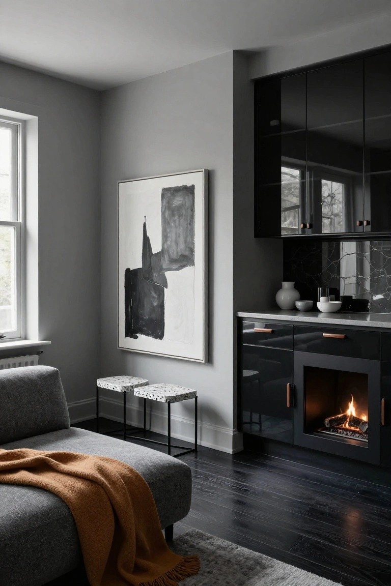 Gray interior room corner featuring glossy black kitchen cabinets with copper handles, integrated gas fireplace, large white-framed abstract black painting on wall, gray sofa with orange throw blanket, two speckled stools, and white ceramic vessels on concrete countertop.