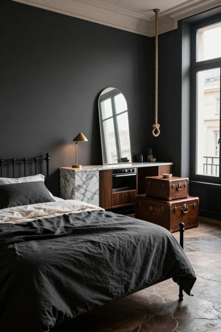 Moody bedroom interior with dark gray walls, black iron bed draped in gray and white linens, two stacked brown leather suitcases beside the bed serving as nightstand, marble-topped wood cabinetry with oven drawer, arched mirror, gold cone lamp, rope hanging from ceiling, and large window with balcony view.