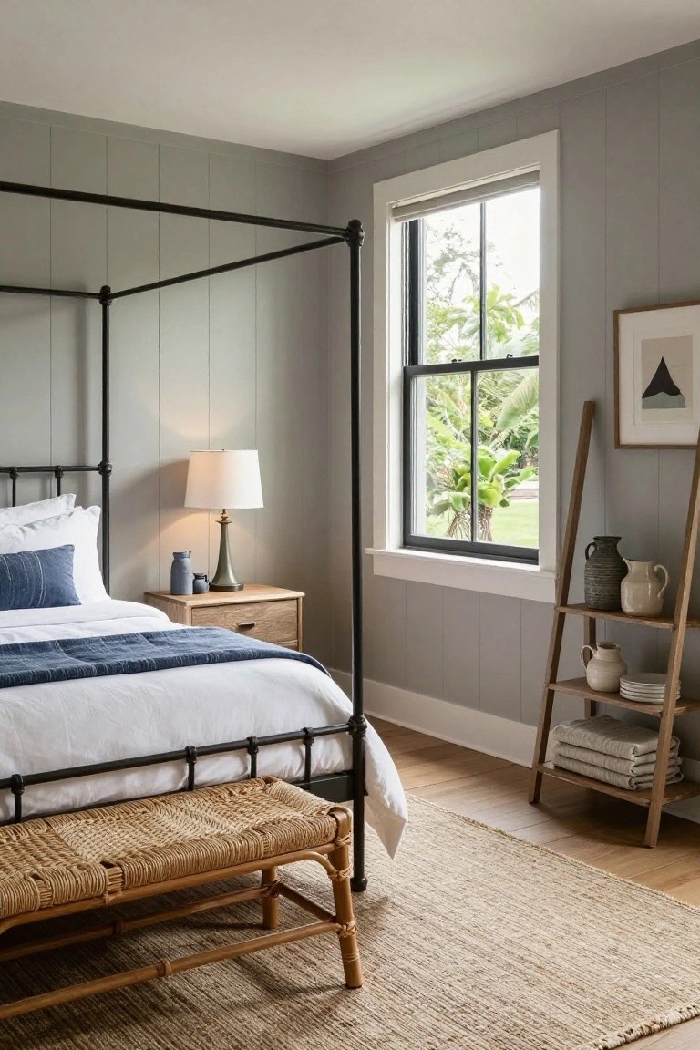 Bedroom with black metal canopy bed frame, white bedding with blue throw, wooden nightstand and lamp, ladder shelf holding ceramics and linens, rattan bench at bed foot, pale gray shiplap walls, and window showing tropical plants outside.
