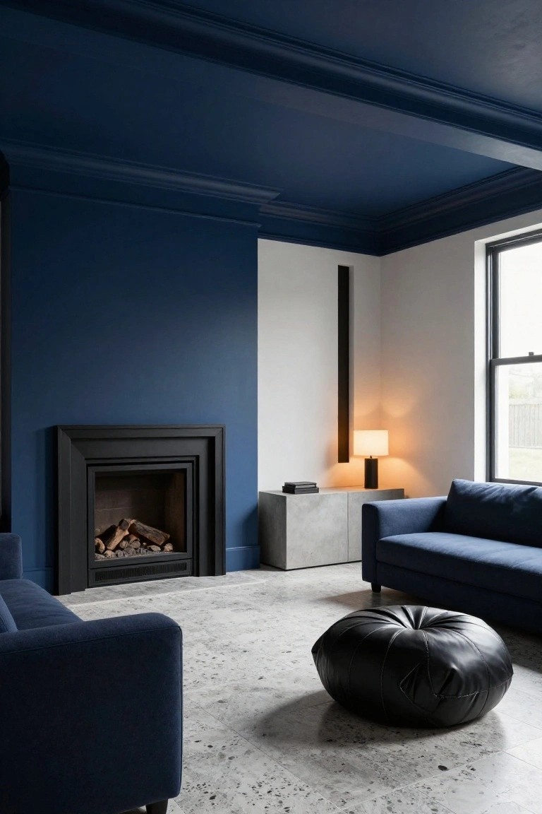 Living room with navy blue ceiling and three walls, white accent wall, black fireplace containing logs, gray concrete cabinet with lamp and books, two navy blue sofas, black leather pouf on speckled gray floor, large window.