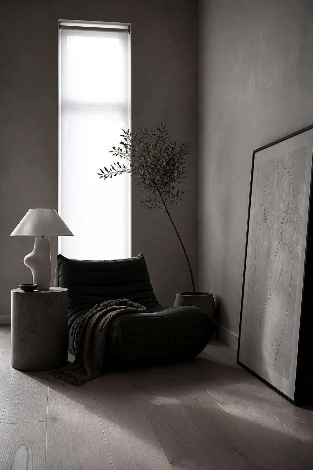 A minimalist living room corner featuring a low black lounge chair beside a tall window, with a concrete side table holding a lamp, a potted tree, and a large framed artwork on the wall.
