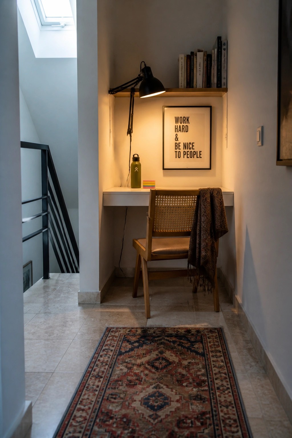 A small white desk with a wooden chair is tucked into a narrow interior alcove beneath a wooden shelf that holds books and a black task lamp.