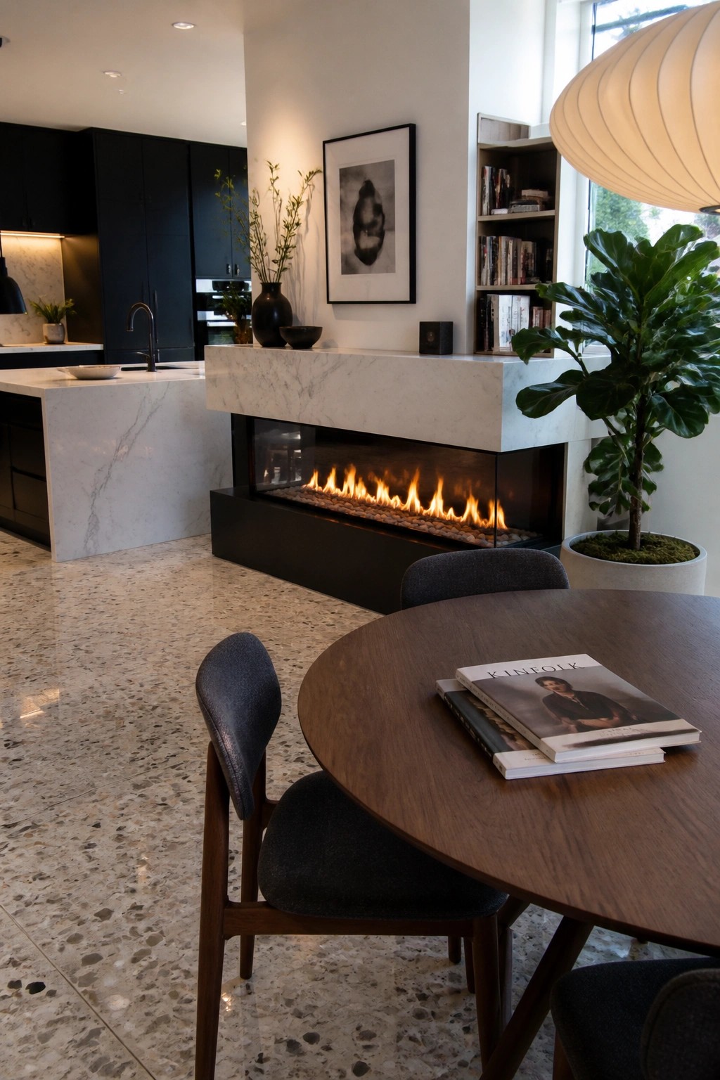 Modern dining area with round wooden table, lit fireplace, and kitchen island.