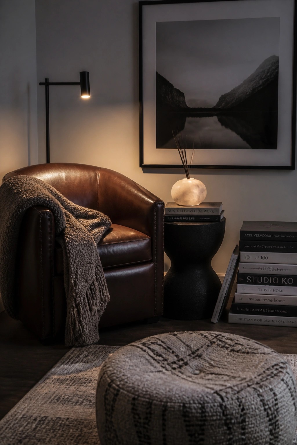 A brown leather armchair with a gray fringed throw blanket sits in a bedroom corner beside a black sculptural side table holding books and a round marble lamp, with a large framed landscape photo above it.