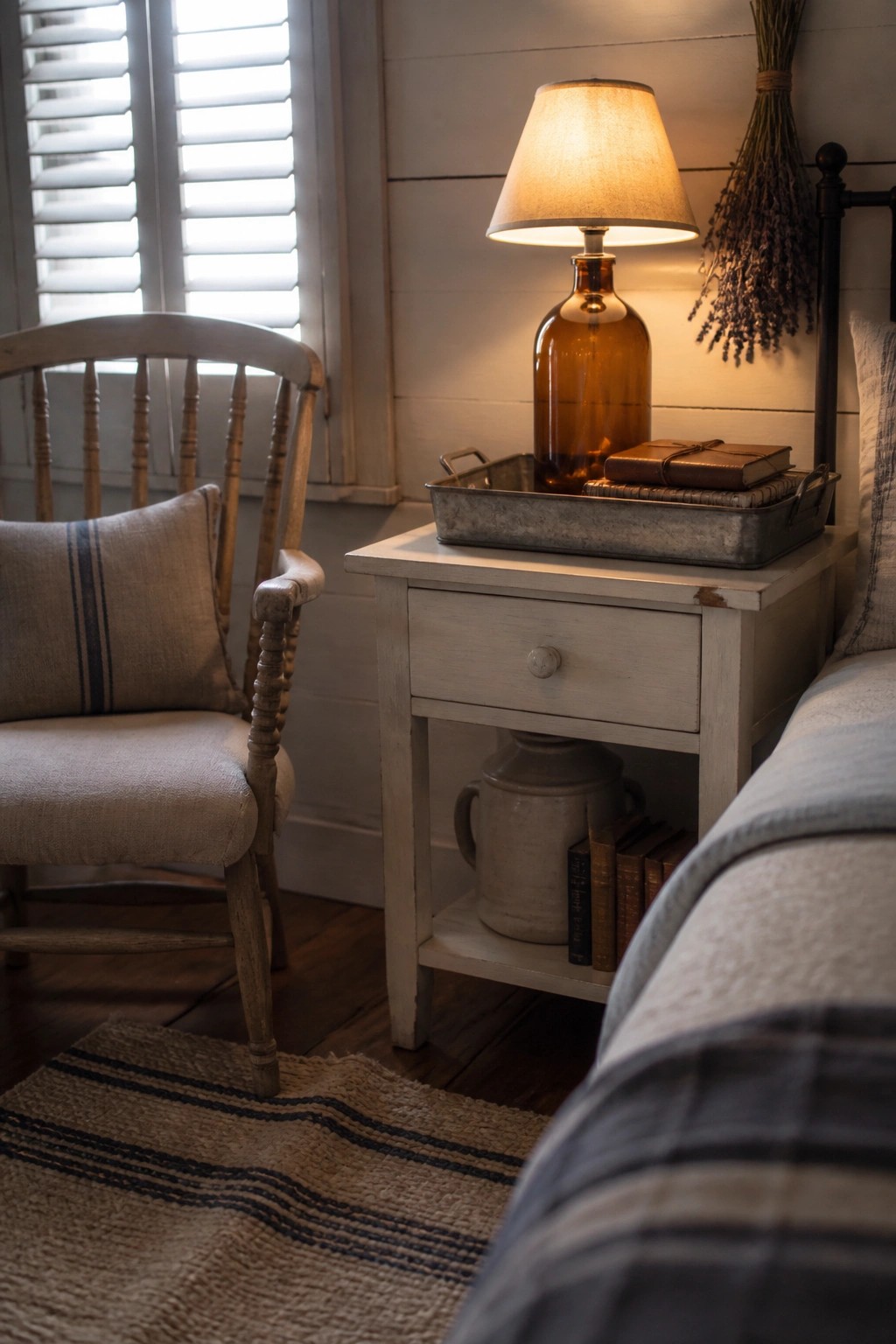 A bedroom corner with a white wooden nightstand holding an amber glass lamp on a metal tray, next to a wooden chair with a striped pillow.