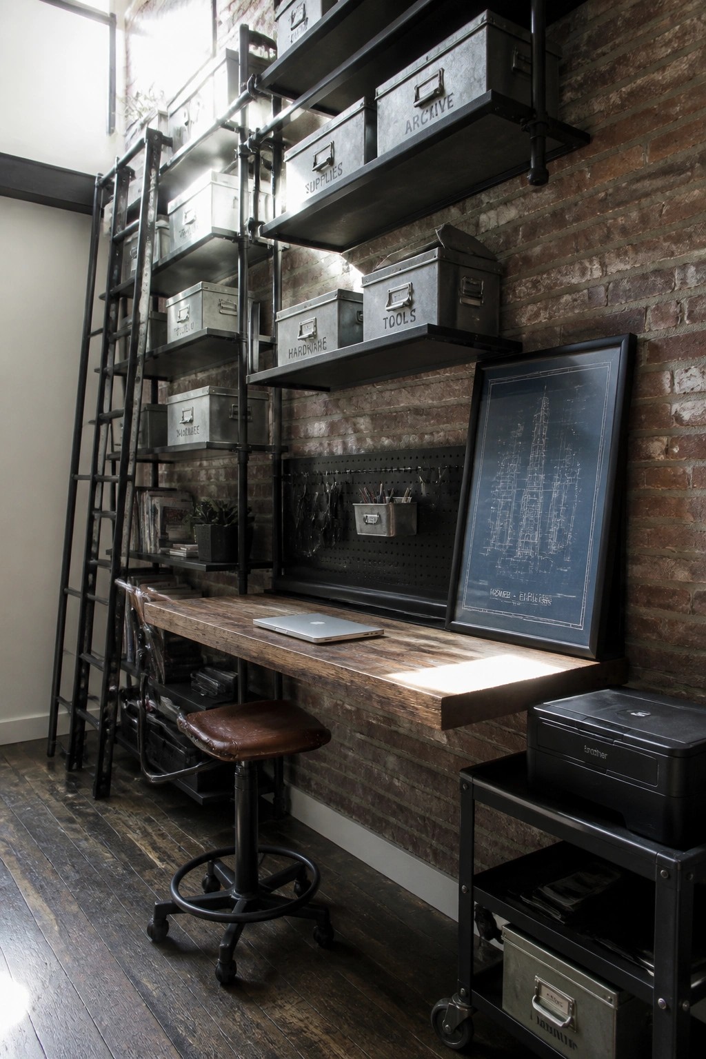 A narrow home office with a long wooden desk against a brick wall, black pipe shelving holding labeled metal bins, a laptop on the desk, and a rolling printer cart nearby.