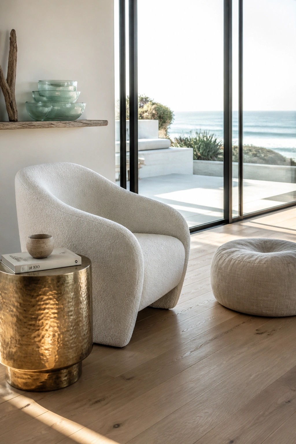 A white textured accent chair next to a round fabric ottoman and a brass side table on light wood flooring, with large glass doors open to the ocean.
