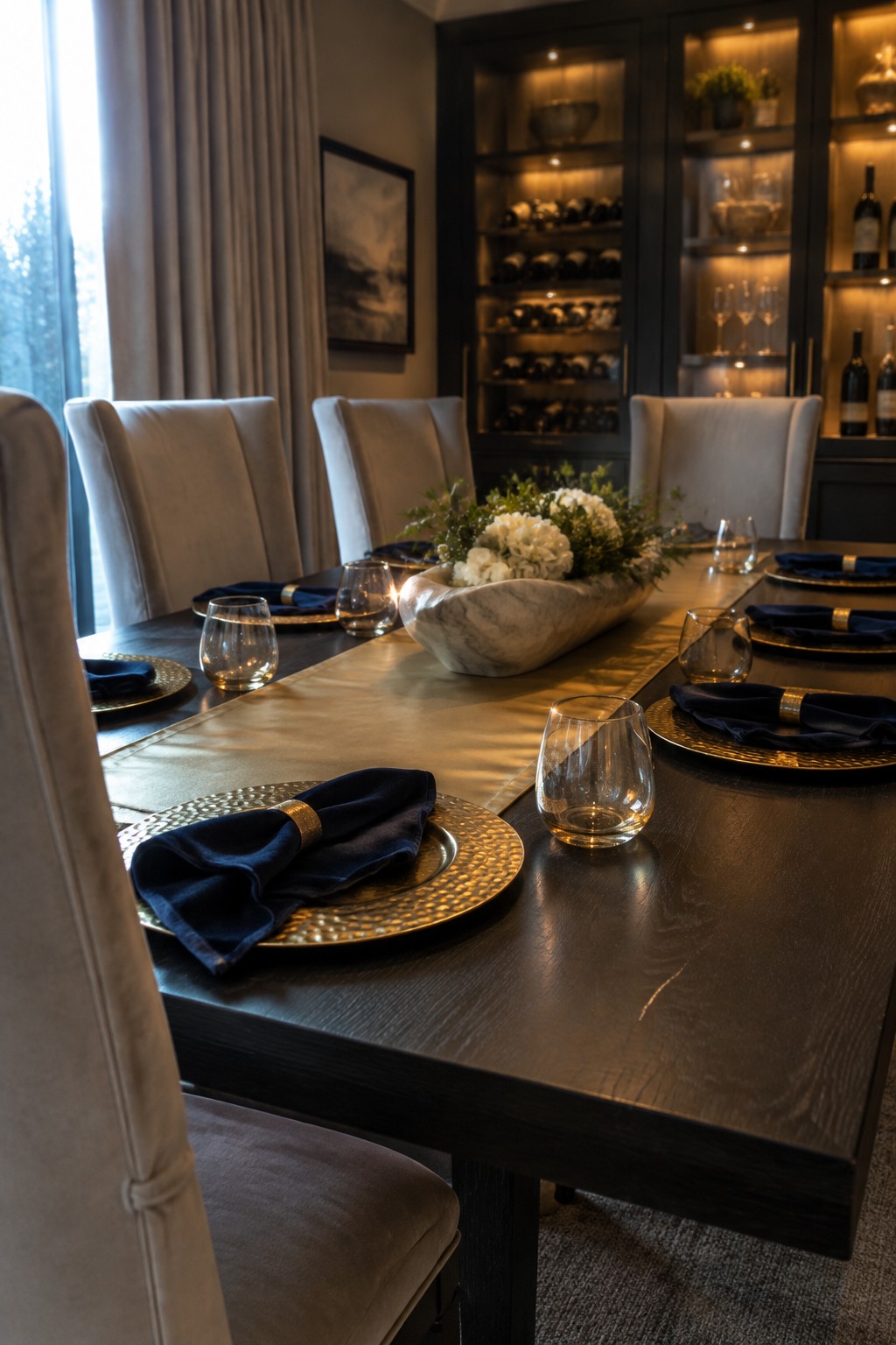 A dark dining table is set with gold chargers, navy napkins, clear glasses, a pale runner, and a white floral centerpiece.