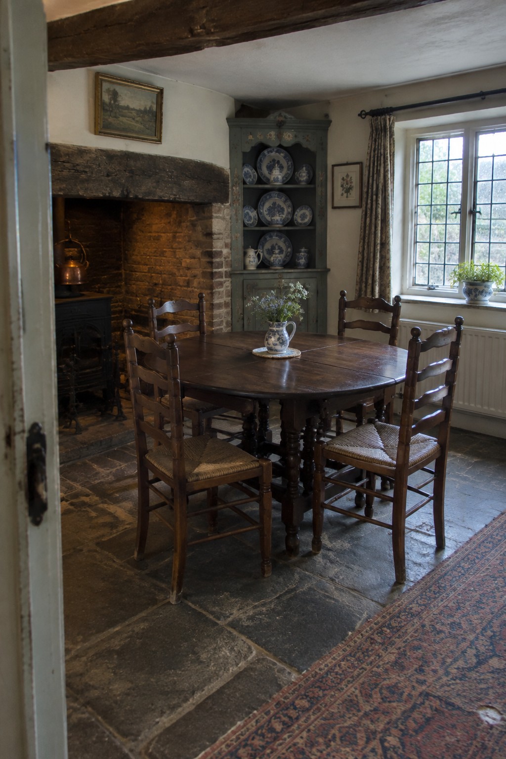 A wooden dining table and ladder-back chairs sit beside a large brick fireplace in a traditional room.