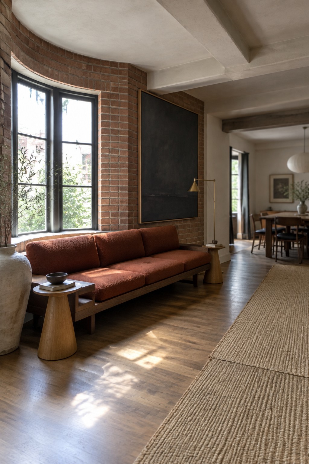 A rust-colored sofa sits beneath a large dark artwork mounted on an exposed brick wall beside tall windows.