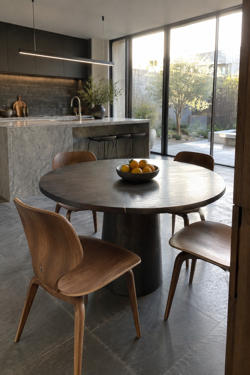 A round dark wood dining table with wooden chairs sits beside a marble kitchen island and floor-to-ceiling windows.