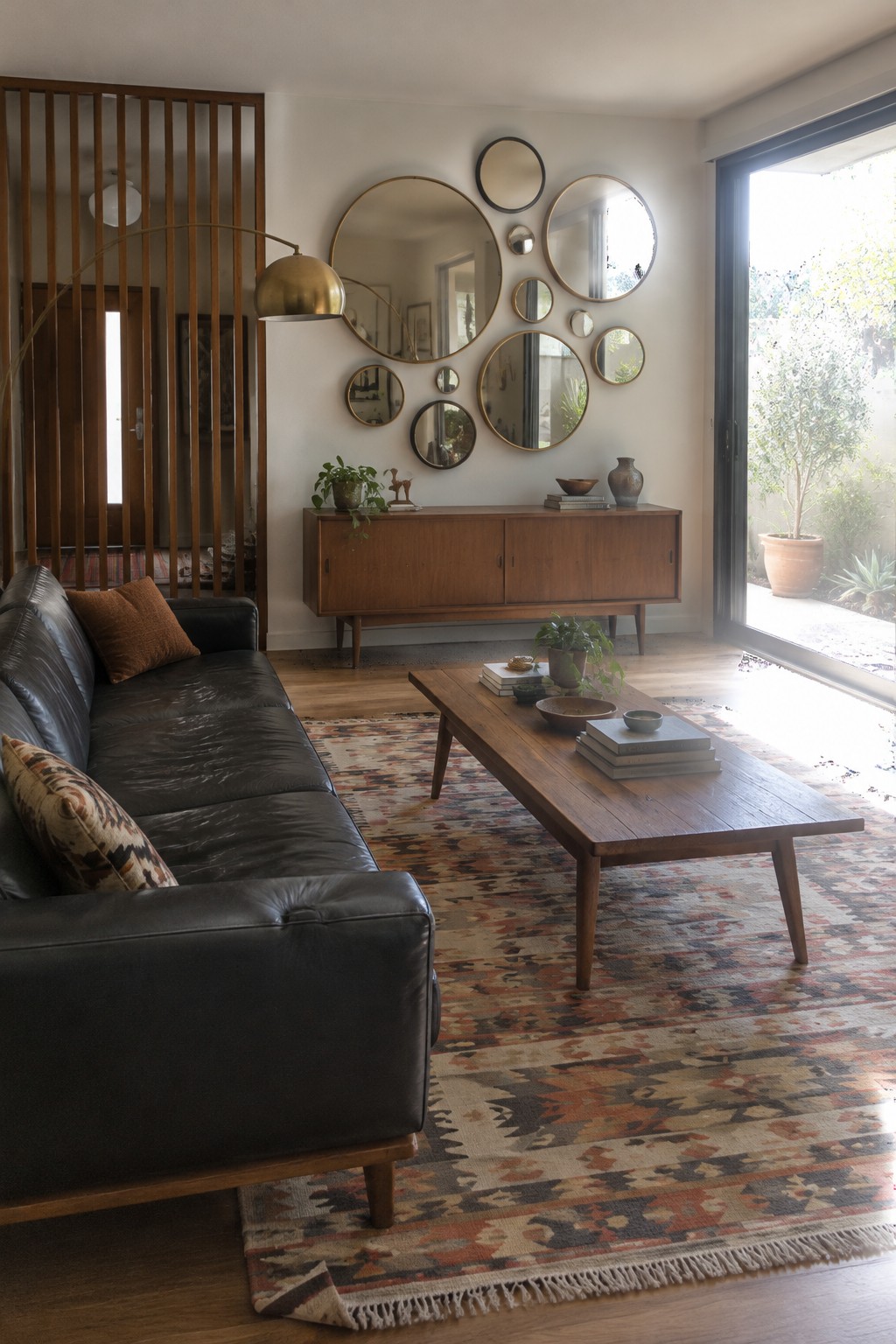 A dark leather sofa sits beside a wooden coffee table on a patterned rug, with a wood sideboard and round mirrors in the living room.