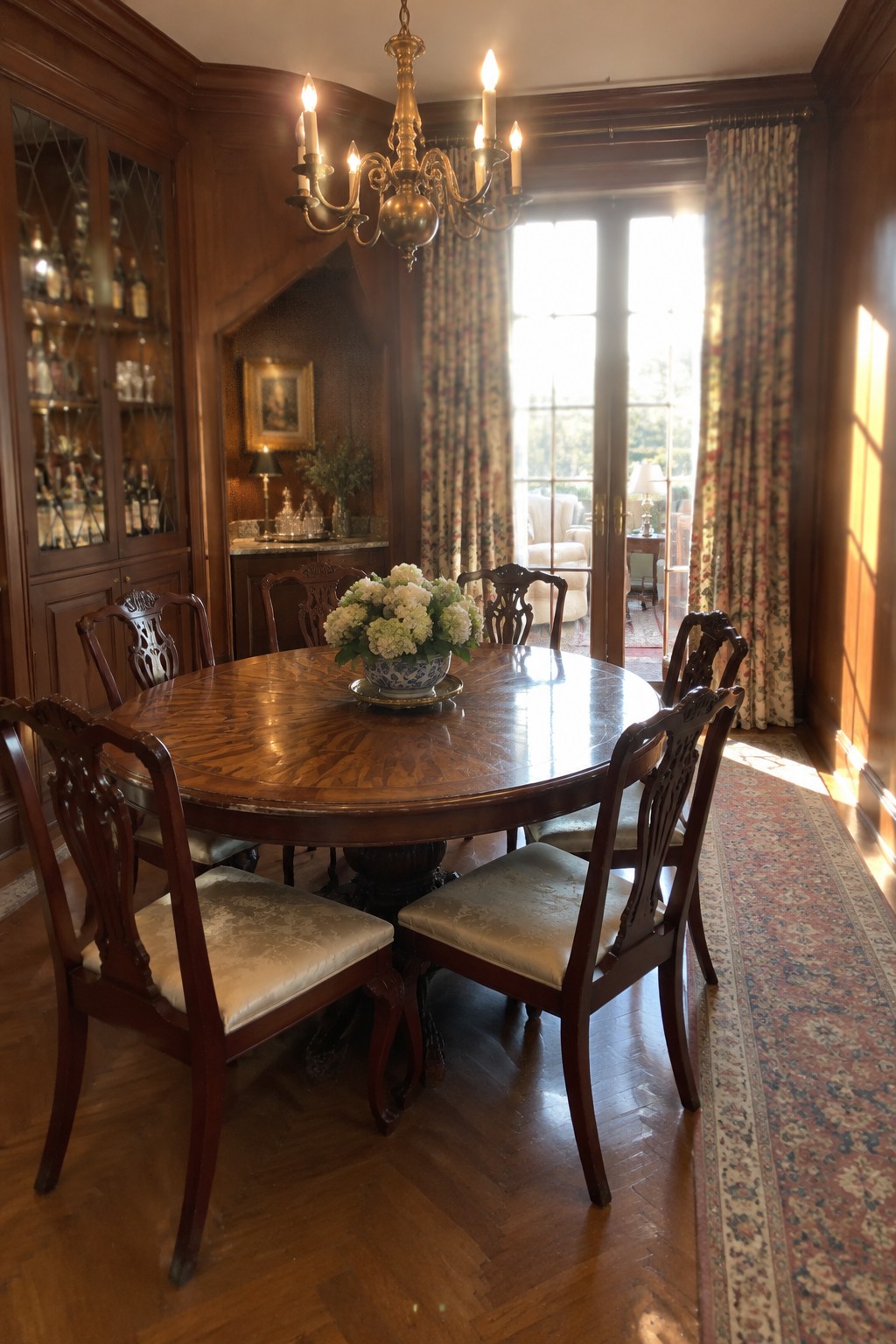 A round wooden dining table and carved chairs sit in a wood-paneled room with a glass-front cabinet and French doors.